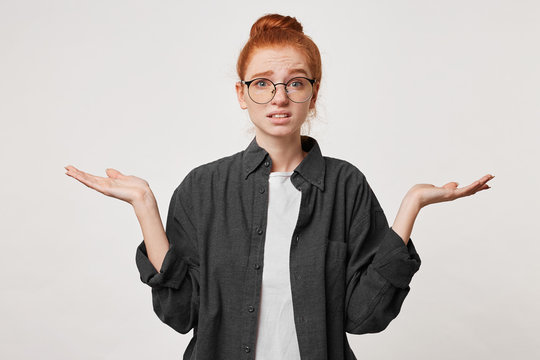 Studio photo of a girl with red hair wearing glasses dressed in a man's black shirt depicts helplessness hopelessness holding hands to the sides with her palms upwards. Confused scared apologizes.
