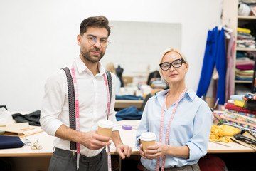 Waist up portrait of two mature tailors looking at camera while posing in atelier holding coffee cups