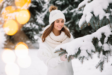 Outdoor close up portrait of young beautiful happy smiling girl wearing white knitted beanie hat, scarf and gloves. Model posing in park with Christmas lights. Winter holidays concept.