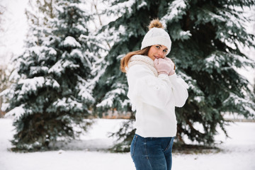 Outdoor close up portrait of young beautiful happy smiling girl wearing white knitted beanie hat, scarf and gloves. Model posing in park with Christmas lights. Winter holidays concept.