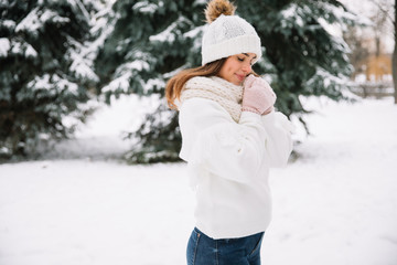 Outdoor close up portrait of young beautiful happy smiling girl wearing white knitted beanie hat, scarf and gloves. Model posing in park with Christmas lights. Winter holidays concept.