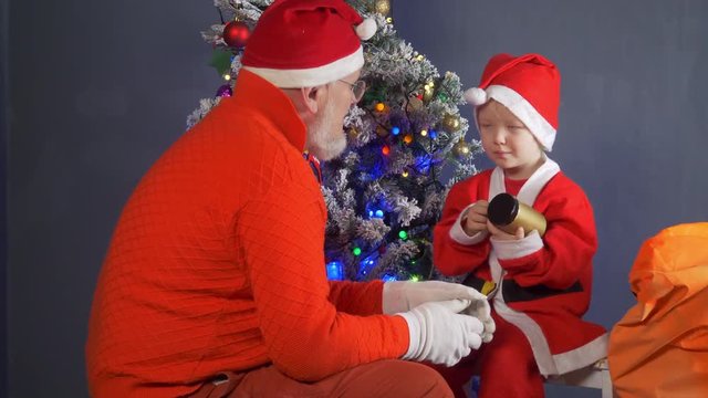 Happy child Dressed in Santa Claus costume, pulls yellow pig toy out of gift box. father in Santa Claus hat helps him against background of Christmas tree in garlands. Symbol of  chinese new year