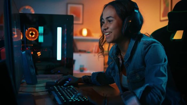 Pretty and Excited Black Gamer Girl in Headphones is Playing and Winning in First-Person Shooter Online Video Game on Her Computer. Room and PC have Colorful Neon Led Lights. Cozy Evening at Home.
