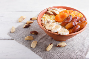 A plate with muesli, banana, dried apricots, dates, Brazil nuts on a white wooden background.