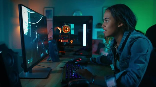 Beautiful and Excited Black Gamer Girl is Playing First-Person Shooter Online Video Game on Her Computer. Room and PC have Colorful Neon Led Lights. Cozy Evening at Home.