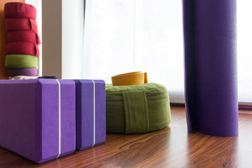 Various yoga props on studio wooden floor. Set of purple blocks, yellow belt on green meditation pouf, standing mat and pile of colorful cushions on background. Wellness, workout activity concepts