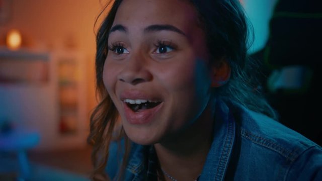 Close Up Portrait of a Beautiful Excited Black Girl Watching an Action Video on a Computer. She Has Dark Hair and Brown Eyes. Screen Adds Reflections to Her Face. Cozy Room is Lit with Warm Light.