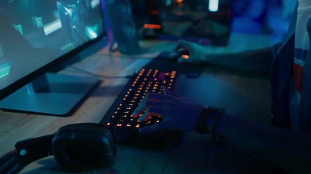 Close Up Hands Shot Showing a Gamer Pushing the Keyboard Buttons while Playing an Online Shooter Video Game. Keyboard Led Lights Change Color in Rainbow Spectrum. Gamer is Wearing a Bracelet.