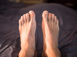 Personal perspective looking down at a young adult male or man's bare feet and toes pointed up laying on a bed with blue comforter bedding below and in the background.