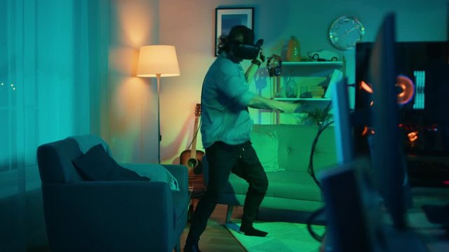 Young Man is Energetically Playing a Virtual Reality Video Game While Wearing a Headset. He's is Standing in a Cozy Room with Warm Neon Lights. He's Swinging Hands, Shooting and Dodging Obstacles.
