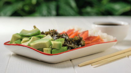 Bowl with rice, avocado, salmon and kelp on a white wooden table against a green tree.