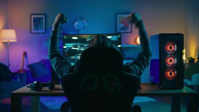 Back Shot of a Gamer Playing and Winning in First-Person Shooter Online Video Game on His Powerful Personal Computer. Room and PC have Colorful Neon Led Lights. Cozy Evening at Home.