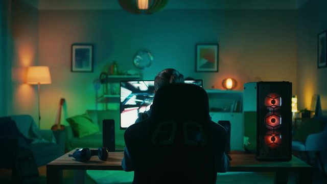 Back Shot of a Gamer Playing and Winning in First-Person Shooter Online Video Game on His Powerful Personal Computer. Room and PC have Colorful Neon Led Lights. Cozy Evening at Home.