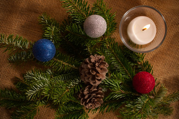 New Year and Christmas decoration with a white burning candle in transparent glass bowl, colorful toys, a branch of a fir tree lying on the brown texture canvas fabric as background. View from above.