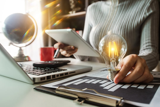 business hand holding lightbulb with using laptop computer and money stack in office. idea saving energy and accounting finance concept  in morning light