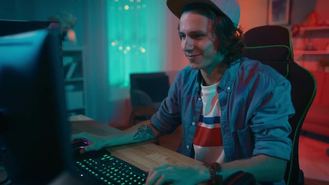 Excited Gamer Playing and Winning in Online Video Game on His Personal Computer. Room and PC have Colorful Warm Neon Led Lights. Young Man is Wearing a Cap. Cozy Evening at Home.