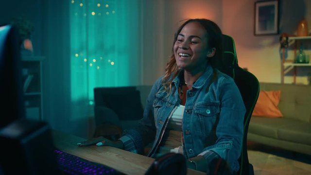 Beautiful Surprised and Excited Young Black Girl Watching Action Video on a Computer. She Has Dark Hair and Brown Eyes. Screen Adds Reflections to Her Face. Cozy Room is Lit with Warm Light.