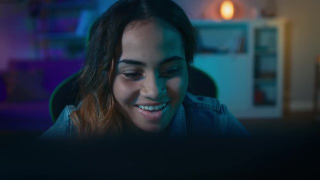 Beautiful Surprised and Excited Young Black Girl Watching Action Video on a Computer. She Has Dark Hair and Brown Eyes. Screen Adds Reflections to Her Face. Cozy Room is Lit with Warm Light.