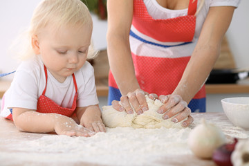 Obraz na płótnie Canvas Little girl and her blonde mom in red aprons playing and laughing while kneading the dough in kitchen. Homemade pastry for bread, pizza or bake cookies. Family fun and cooking concept