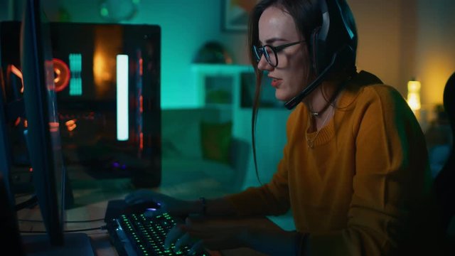 Excited Gamer Girl in Headset with a Mic Playing Online Video Game on Her Personal Computer. She Talks to Other Players. Room and PC have Colorful Warm Neon Led Lights. Cozy Evening at Home.
