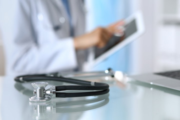 Stethoscope lying on glass desk with laptop computer at busy physician background. Medicine or pharmacy concept. Medical tools at doctor working table