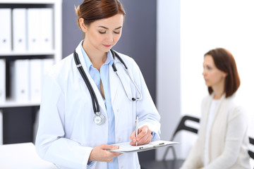 Doctor woman at work. Portrait of female physician filling up medical form while standing near reception desk at clinic or emergency hospital. Patient woman sitting at the background. Medicine concept