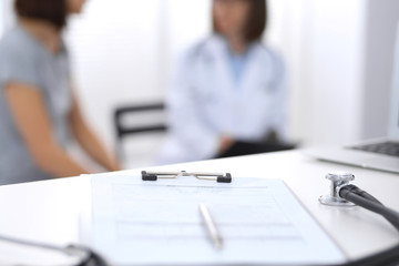 Stethoscope, clipboard with medical form lying on hospital reception desk with laptop computer and busy doctor and patient communicating at the background. Medical tools at doctor working table