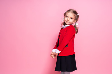 Adorable little girl in red school jacket and black dress looking at camera while standing on pink background