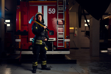 Full-length photo of firefighter in protective helmet with hammer looking at camera against background of fire engine