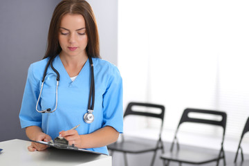 Doctor woman at work. Portrait of female physician filling up medical form while standing near reception desk at clinic or emergency hospital. Medicine and healthcare concept