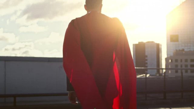 Businessman Superhero WIth Red Cape Blowing in the Wind Walks on the Roof of a Skyscraper, Looking into the Sunset, Ready to Save the Day. Following Back View Slow Motion Shot.