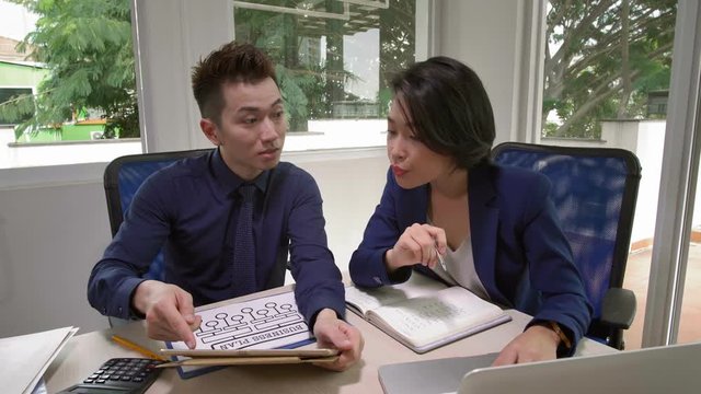 Medium shot of young Asian businessman and businesswoman sitting together at working table, looking at tablet in hands of businessman and discussing business plan