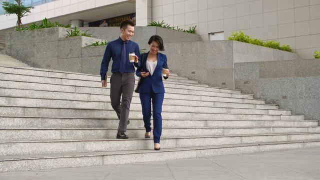 Wide shot of male and female business colleagues, walking together down stairs, holding paper cups with coffee and talking