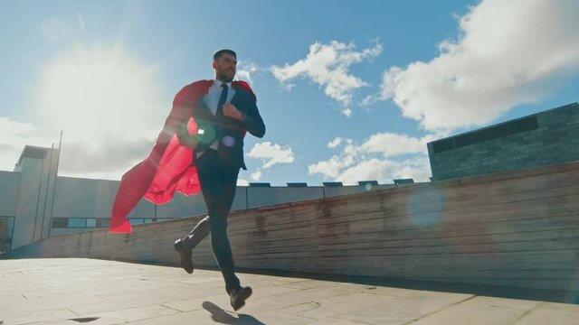Businessman Superman WIth Red Cape Blowing in the Wind Runs on the Roof of a Skyscraper, Ready to Save the Day. Following Back View Slow Motion Shot.