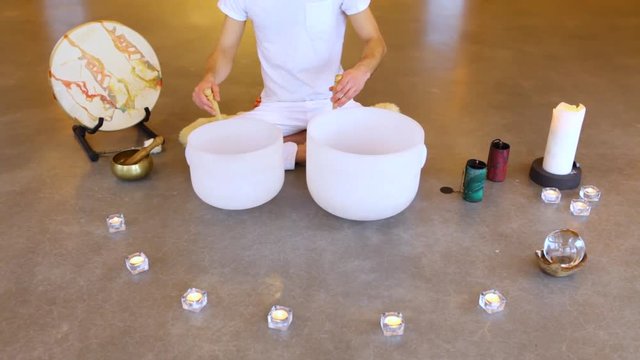 Man playing crystal bowls healing music. Travelling up of a young man dressed in white, sitting in indian surrounded by instruments and playing his two crystal bowls as part of a meditative concert