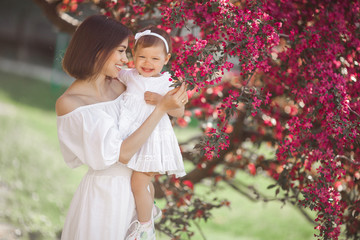 Portrait of young beautiful mother with her little baby girl. Close up still of loving family. Attractive woman holding her child in pink flowers and smiling.