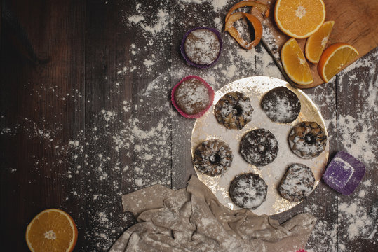 A photo of tasty cakes sprinkled with sweet powder and chocolate on a wooden brown table. Oranges and instruments  for prepare cakes. Composition with cupcakes on the table