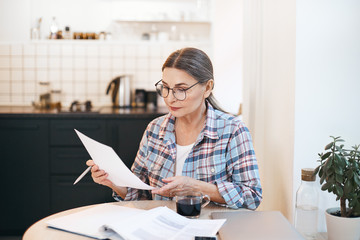 Stylish serious elderly Caucasian woman in spectacles and plaid shirt studying paper in her hand...