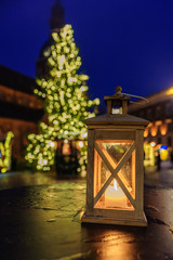 Blurred view of fir tree with glowing Christmas lights near the table with Christmas lantern. Festive mood. Outdoors composition. Christmas and New Year concept.