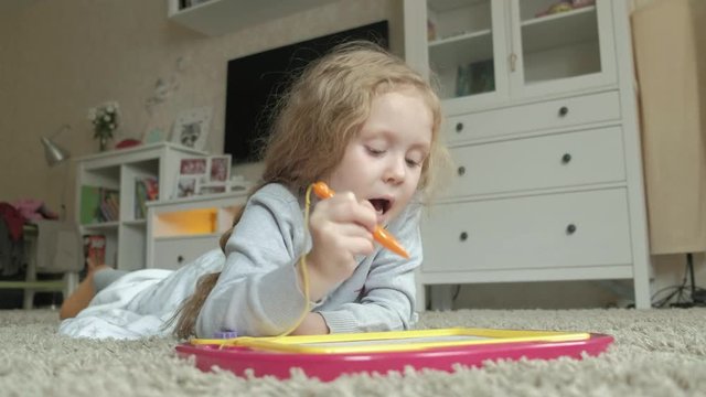 A little girl with red wavy hair lies on the floor and draws on a magnetic board. The concept of the educational process.