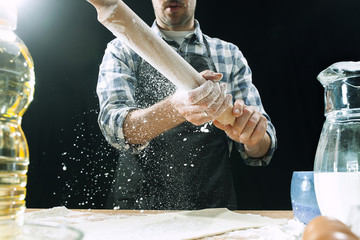Professional male cook sprinkles dough with flour, preapares or bakes bread or pasta at kitchen table, has dirty uniform, isolated over black chalk background. Baking concept
