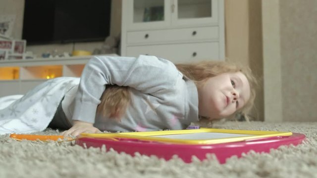 A little girl with red wavy hair lies on the floor and draws on a magnetic board. The concept of the educational process. close-up