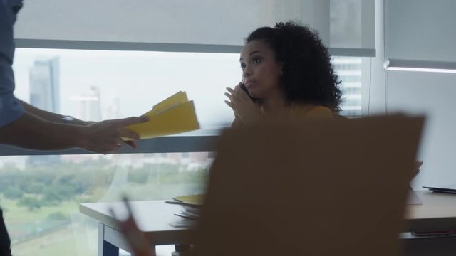 Young business woman working in office. Black businesswoman talking on mobile telephone. African american manager or secretary using smartphone for cell phone conversation and colleague giving her pap