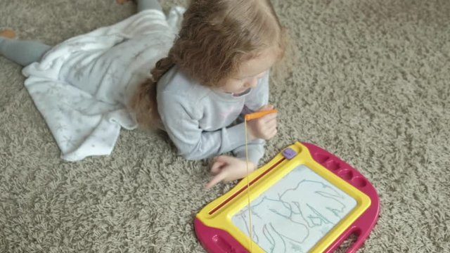 A little girl with red wavy hair lies on the floor and draws on a magnetic board. The concept of the educational process.