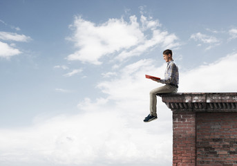 Man on roof edge reading book and cloudscape at background