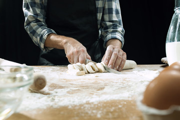 Professional male cook sprinkles dough with flour, preapares or bakes bread or pasta at kitchen table, has dirty uniform, isolated over black chalk background. Baking concept