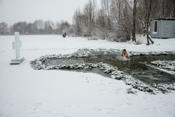 A man bathes in an ice hole. The ice cross is installed at the hole. Behind the cross, fishermen catch fish. Hole for ritual washing. Ukraine. Belogorodka. January 19, 2018