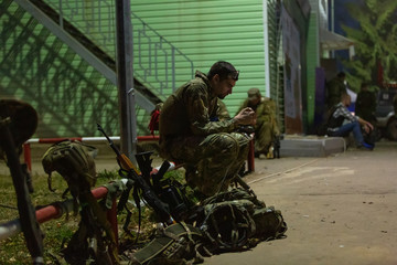 Side view serious soldier writing message in phone while wearing in uniform and surrounded by weapons. Technology and military concept