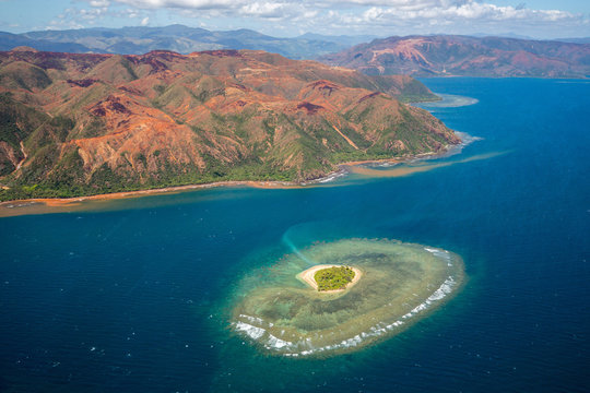 A small atoll islet with heart shaped coral reef off the east coast of Grande Terre island of New Caledonia, French overseas collectivity. Red green mountains hills full of nickel ore near Nakety.
