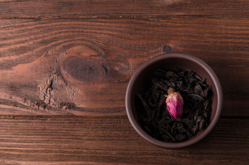 The tea cup, with a loose dried black tea leaves and a tea rose bud, is on the wooden table.